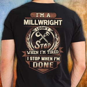 Op de afbeelding: Zwart T-shirt met een bruin en beige opdruk. De opdruk bevat de tekst "I'M A MILLWRIGHT I DON'T STOP WHEN I'M TIRED I STOP WHEN I'M DONE" en een gekruist ontwerp van een moersleutel en schuifmaat.