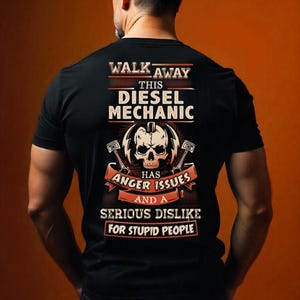 Op de afbeelding: Zwart t-shirt met een grafisch ontwerp met een schedel, zuigers en de tekst "WALK AWAY THIS DIESEL MECHANIC HAS ANGER ISSUES AND A SERIOUS DISLIKE FOR STUPID PEOPLE."