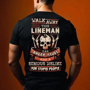 May include: Black t-shirt with a graphic design. The design includes the text "WALK AWAY THIS LINEMAN" above a skull and lightning bolts, with the text "HAS ANGER ISSUES AND A SERIOUS DISLIKE FOR STUPID PEOPLE" below.