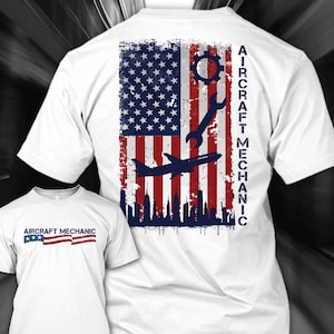 Può includere: T-shirt bianca con un design a bandiera americana usurata con ingranaggio, chiave inglese e silhouette di aereo. Il testo "AIRCRAFT MECHANIC" è stampato verticalmente sul retro. Una versione più piccola del design è sul davanti.
