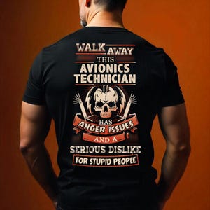 Op de afbeelding: Zwart t-shirt met de tekst "WALK AWAY THIS AVIONICS TECHNICIAN HAS ANGER ISSUES AND A SERIOUS DISLIKE FOR STUPID PEOPLE." Het ontwerp bevat een schedel en bliksemschichten.