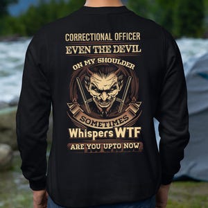 May include: Black sweatshirt with a graphic design on the back. The design features a devil face with crossed weapons, and text that reads "CORRECTIONAL OFFICER EVEN THE DEVIL ON MY SHOULDER SOMETIMES Whispers WTF ARE YOU UPTO NOW."