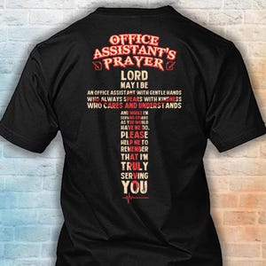 May include: A black short-sleeved t-shirt featuring the text "OFFICE ASSISTANT'S PRAYER" in red and white at the top. Below, a cross-shaped graphic contains the prayer text in white, with the word "PLEASE" highlighted in red letters.