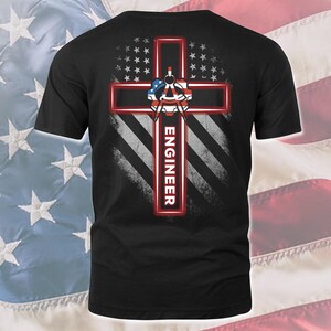 May include: Black t-shirt featuring a red cross design with the word "ENGINEER" in white. The cross is overlaid on a distressed American flag design, with a compass and protractor graphic at the center. The shirt has short sleeves.