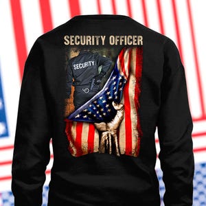 Könnte beinhalten: Schwarzes Sweatshirt mit einer Grafik der amerikanischen Flagge, die von einer Hand gehalten wird, und einem Sicherheitsbeamten in Uniform. Die Worte "SECURITY OFFICER" sind oben auf dem Design aufgedruckt.