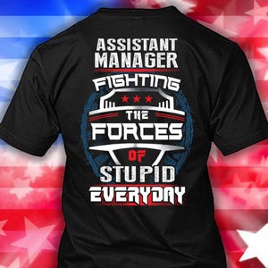 Pode incluir: Camiseta preta com o texto "ASSISTANT MANAGER FIGHTING THE FORCES OF STUPID EVERYDAY" em letras brancas e vermelhas. O design inclui um gráfico em forma de escudo com estrelas e uma borda azul.