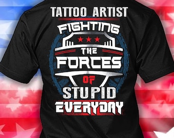 Może przedstawiać: Czarna koszulka z napisem "TATTOO ARTIST FIGHTING THE FORCES OF STUPID EVERYDAY" w białych i czerwonych literach. Projekt zawiera niebiesko-białą okrągłą grafikę z gwiazdami.