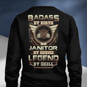 Puede incluir: Sudadera negra con un diseño gráfico. El diseño presenta las palabras "BADASS BY BIRTH", "JANITOR BY CHOICE" y "LEGEND BY SKILL" en una fuente estilizada. El diseño también incluye un emblema circular con alas y herramientas de limpieza.