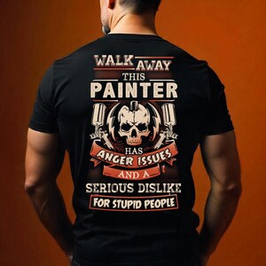 Könnte beinhalten: Ein schwarzes T-Shirt mit einer Grafik auf der Rückseite, die einen Totenkopf, zwei Farbsprühpistolen und den Text zeigt: WALK AWAY THIS PAINTER HAS ANGER ISSUES AND A SERIOUS DISLIKE FOR STUPID PEOPLE. Das Design verwendet weiße, orange und schwarze Töne.