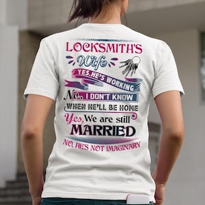 Puede incluir: Camiseta blanca con el texto "LOCKSMITH'S Wife" y otras frases sobre la esposa de un cerrajero. El diseño incluye llaves y pancartas decorativas en varios colores.