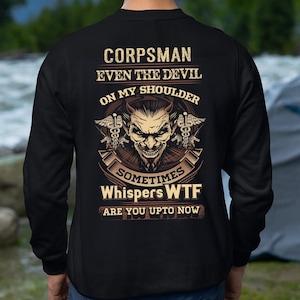 Puede incluir: Sudadera negra con un diseño gráfico en la espalda. El diseño presenta el texto "CORPSMAN EVEN THE DEVIL ON MY SHOULDER" sobre una cara de diablo y el texto "SOMETIMES Whispers WTF ARE YOU UPTO NOW".