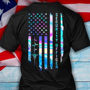 Könnte beinhalten: Schwarzes T-Shirt mit einem holografischen Design der amerikanischen Flagge. Die Flagge zeigt Sterne und Streifen in Blau, Lila und Rosa. Der Text "SECURITY OFFICER" ist vertikal auf der rechten Seite der Flagge aufgedruckt.