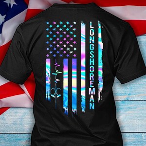 Puede incluir: Camiseta negra con un diseño colorido de la bandera estadounidense. La bandera presenta estrellas y rayas en tonos azules, rosas y morados, con el texto "LONGSHOREMAN" y un gráfico de latido cardíaco y anzuelo.
