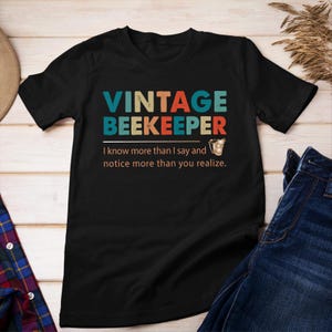 May include: Black t-shirt with a vintage-style graphic that says "Vintage Beekeeper" in colorful letters. The graphic also includes a small image of a honey dipper. The text below the graphic says "I know more than I say and notice more than you realize."
