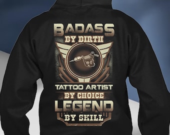 Może przedstawiać: Czarna bluza z kapturem z graficznym wzorem maszynki do tatuażu. Tekst "BADASS BY BIRTH, TATTOO ARTIST BY CHOICE, LEGEND BY SKILL" jest w brązowej, postarzanej czcionce. Projekt ma wygląd vintage.