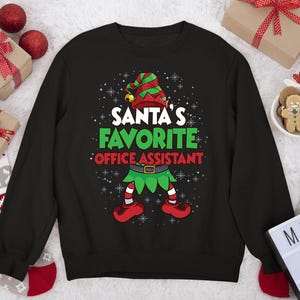 May include: Black sweatshirt with a festive graphic. The graphic features an elf design with the text "SANTA'S FAVORITE OFFICE ASSISTANT" in green and red. The elf is wearing a green and red hat and shoes.