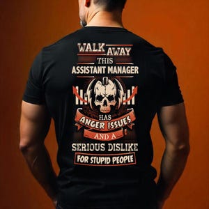 Könnte beinhalten: Schwarzes T-Shirt mit einem grafischen Design auf der Rückseite. Das Design enthält den Text "WALK AWAY THIS ASSISTANT MANAGER HAS ANGER ISSUES AND A SERIOUS DISLIKE FOR STUPID PEOPLE" mit einem Totenkopf und dekorativen Elementen.