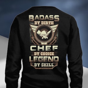 May include: Black long-sleeve sweatshirt with a graphic design for chefs. The design includes the words "BADASS BY BIRTH", "CHEF BY CHOICE", and "LEGEND BY SKILL", along with a chef's hat and crossed knives.