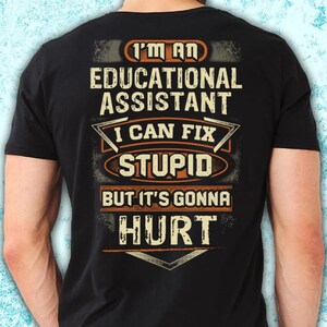 May include: Black t-shirt featuring the text "I'M AN EDUCATIONAL ASSISTANT I CAN FIX STUPID BUT IT'S GONNA HURT" in white, orange, and grey. The design has a distressed, vintage appearance.