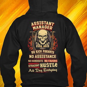 Op de afbeelding: Zwarte hoodie met de tekst "ASSISTANT MANAGER" en een schedelafbeelding. Daaronder staat de tekst "NO RICH PARENTS NO ASSISTANCE NO HANDOUTS NO FAVORS STRAIGHT HUSTLE Au Day Everyday."