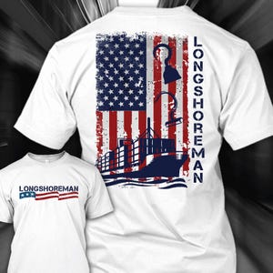 Puede incluir: Camiseta blanca con un diseño desgastado de la bandera estadounidense, un barco y un ancla. El texto "LONGSHOREMAN" está impreso verticalmente en la espalda y horizontalmente en la parte delantera. El diseño utiliza los colores rojo, blanco y azul.