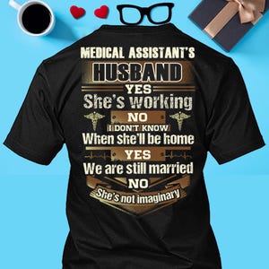 Puede incluir: Camiseta negra con el texto "MEDICAL ASSISTANT'S HUSBAND". La camiseta presenta un diseño humorístico con frases como "She's working", "When she'll be home" y "We are still married". El texto es en blanco y dorado.
