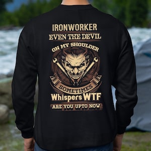 Puede incluir: Sudadera negra con el texto "IRONWORKER EVEN THE DEVIL ON MY SHOULDER SOMETIMES WHISPERS WTF ARE YOU UPTO NOW" y un gráfico de diablo con llaves cruzadas. El diseño es en tonos marrones y beige.