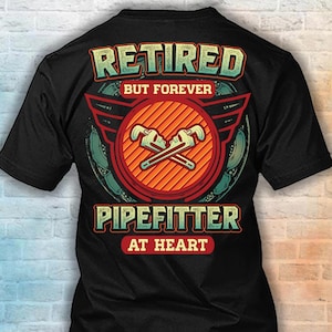 May include: A black short-sleeve t-shirt featuring a graphic on the back with the text "RETIRED BUT FOREVER PIPEFITTER AT HEART". The design includes a central orange circle with two crossed pipe wrenches, surrounded by red wings and teal accents.