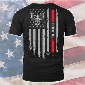 以下が含まれることがあります： アメリカのぼろぼろの旗のデザインが施された黒いTシャツ。旗の中央にはハクトウワシがいて、赤いストライプに「TRUCKER」という文字が縦に書かれています。