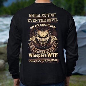 Op de afbeelding: Zwart sweatshirt met een grafisch ontwerp met een duivelsgezicht en de tekst "MEDICAL ASSISTANT EVEN THE DEVIL ON MY SHOULDER SOMETIMES WHISPERS WTF ARE YOU UPTO NOW". Het ontwerp is in bruin- en beigetinten.