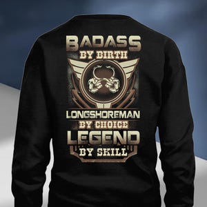 Puede incluir: Sudadera negra con un diseño gráfico. El diseño incluye las palabras "BADASS BY BIRTH", "LONGSHOREMAN BY CHOICE" y "LEGEND BY SKILL". El gráfico también presenta un emblema circular con ganchos cruzados y alas.