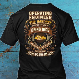 Może przedstawiać: Czarna koszulka z grafiką dla inżyniera eksploatacji. Projekt zawiera tekst "OPERATING ENGINEER" i "THE HARDEST PART OF MY JOB IS BEING NICE". Widoczny jest również żółty pojazd budowlany.