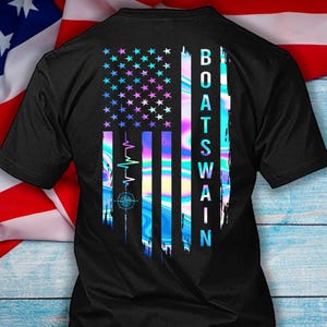 May include: Black t-shirt featuring a stylized American flag design with a vibrant, iridescent color scheme. The flag incorporates stars and stripes, with the word "BOATSWAIN" printed vertically on the right side. A compass and heartbeat graphic are also included.