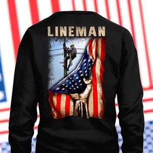 Puede incluir: Sudadera negra con un gráfico de un liniero trabajando en un poste de energía, con un diseño de la bandera estadounidense. La palabra "LINEMAN" está impresa en la parte superior. El diseño incorpora los colores rojo, blanco y azul.