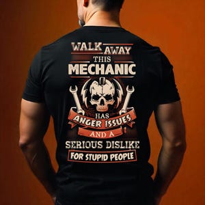Op de afbeelding: Zwart T-shirt met een mechanisch thema. Het ontwerp bevat een schedel, moersleutels en de tekst "WALK AWAY THIS MECHANIC HAS ANGER ISSUES AND A SERIOUS DISLIKE FOR STUPID PEOPLE."