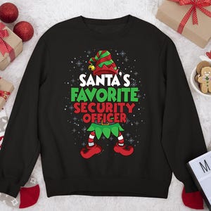 Könnte beinhalten: Schwarzes Sweatshirt mit dem Text "Santa's Favorite Security Officer" in Weiß, Grün und Rot. Das Design umfasst einen Elfenhut, einen Gürtel und Schuhe. Schneeflocken umgeben den Text und das Design und schaffen ein festliches Urlaubsthema.
