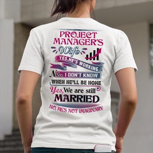 May include: White t-shirt with text in pink and blue. The text reads "PROJECT MANAGER'S Wife YES.HE'S WORKING No, I DON'T KNOW WHEN HE'LL BE HOME Yes, We are still MARRIED No, HE'S NOT IMAGINARY."