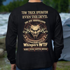 May include: Black sweatshirt with a graphic design featuring a devil face, tow trucks, and text that reads "TOW TRUCK OPERATOR EVEN THE DEVIL ON MY SHOULDER SOMETIMES Whispers WTF ARE YOU UPTO NOW". The design is in shades of brown and tan.