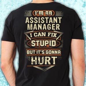 May include: Black t-shirt featuring the text "I'M AN ASSISTANT MANAGER I CAN FIX STUPID BUT IT'S GONNA HURT" in white and orange. The design has a worn, vintage aesthetic.