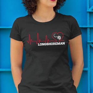 Puede incluir: Camiseta negra con un diseño de línea de latido cardíaco rojo, un corazón y un gráfico de anzuelo. La palabra "LONGSHOREMAN" está impresa en blanco. La camiseta es de manga corta.