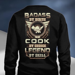 May include: Black sweatshirt with a graphic design. The design features the words "BADASS BY BIRTH", "COOK BY CHOICE", and "LEGEND BY SKILL" in a brown, textured font. A chef's hat and crossed knives are in the center.