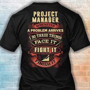 May include: A black short-sleeved t-shirt featuring a graphic design with the text: PROJECT MANAGER WHENEVER A PROBLEM ARRIVES I DO THREE THINGS FACE IT FIGHT IT FINISH IT. The design includes a rising bar graph icon and decorative red banners.