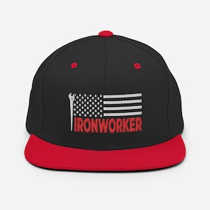 Op de afbeelding: Zwarte en rode baseballpet met een witte Amerikaanse vlag en de tekst "IRONWORKER" geborduurd op de voorkant.