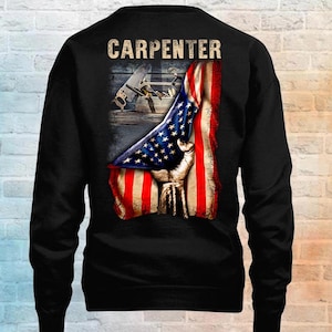 May include: A black long-sleeve sweatshirt featuring a graphic on the back. The graphic displays the word CARPENTER in a tan font above an image of a hand pulling back a United States flag to reveal a wooden surface with various carpentry tools.