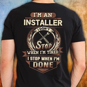 Könnte beinhalten: Ein schwarzes kurzärmeliges T-Shirt mit einem Grafikdruck auf der Rückseite. Der Text lautet I'M AN INSTALLER I DON'T STOP WHEN I'M TIRED I STOP WHEN I'M DONE. Das Design enthält eine zentrale Abbildung von zwei gekreuzten Schraubenschlüsseln.