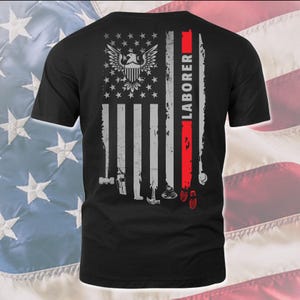 Puede incluir: Camiseta negra con un diseño de bandera estadounidense desgastada. La bandera tiene un águila calva en el centro y la palabra "LABORER" escrita verticalmente en el lado derecho. Hay herramientas y huellas de pies en la parte inferior de la bandera.