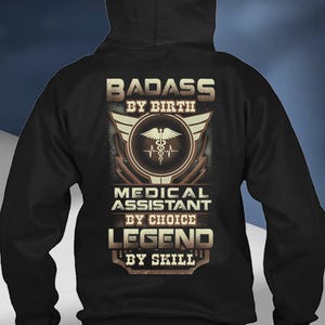 Op de afbeelding: Zwarte hoodie met een grafisch ontwerp. Het ontwerp bevat de tekst "BADASS BY BIRTH, MEDICAL ASSISTANT BY CHOICE, LEGEND BY SKILL" in een distressed, vintage stijl. Een medisch symbool staat in het midden van het ontwerp.