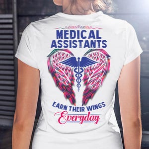 Op de afbeelding: Wit T-shirt met een grafisch ontwerp voor doktersassistenten. Het ontwerp toont de tekst "MEDICAL ASSISTANTS" boven een paar gestileerde vleugels in roze, rood en groen, met een medisch symbool in het midden. De tekst "EARN THEIR WINGS Everyday" staat eronder.