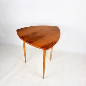 Puede incluir: Una mesa auxiliar triangular de estilo moderno de mediados de siglo con un rico y cálido acabado de vetas de madera. La mesa tiene tres patas delgadas y cónicas. La mesa mide aproximadamente 76 cm de ancho.