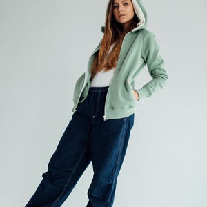 May include: A light green hooded sweatshirt with white fur lining the hood and ears. The sweatshirt has a zipper closure and is worn with dark blue jeans.
